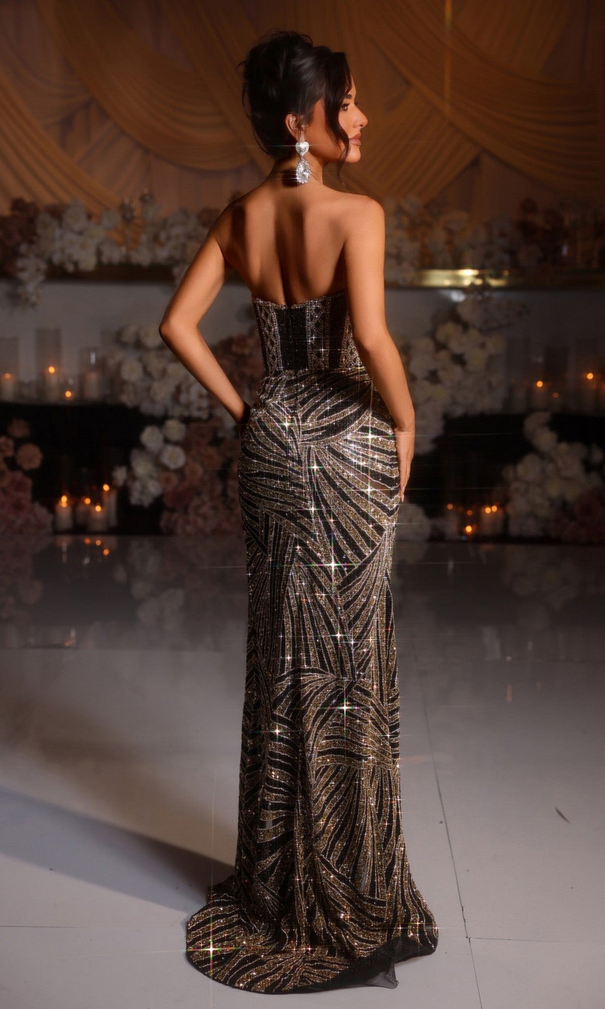 A woman stands indoors with her back to the camera, wearing the strapless, floor-length Portia and Scarlett PS25469C beaded prom dress with a geometric pattern. Floral arrangements and candles decorate the background.